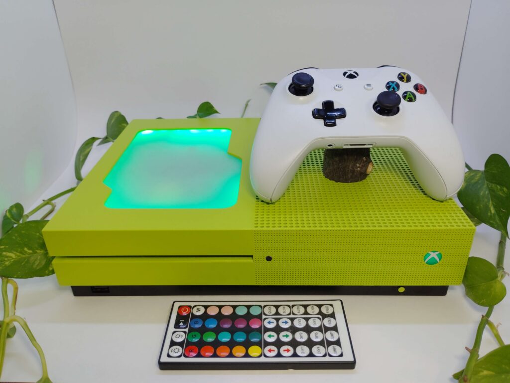 neon green xbox on display with plants surrounding. has a plexiglass window, a custom green led power button and a matching white controller
